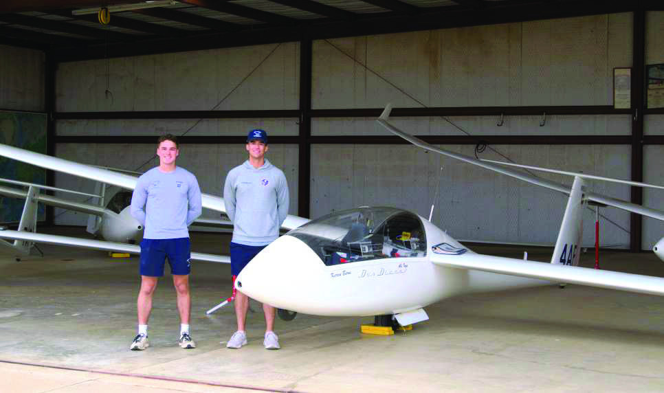 Instructors Matt Withers (left) and Creed Thompson (right), part of the USAF Academy’s 94th line training squadron, are standing beside a TG15A training glider inside a hangar, where aircraft are stored and maintained between flights. (Photo by Leon Szaniszlo)